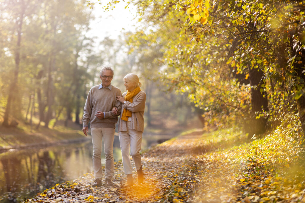Older couple walking together on a wooded trail near a pond.